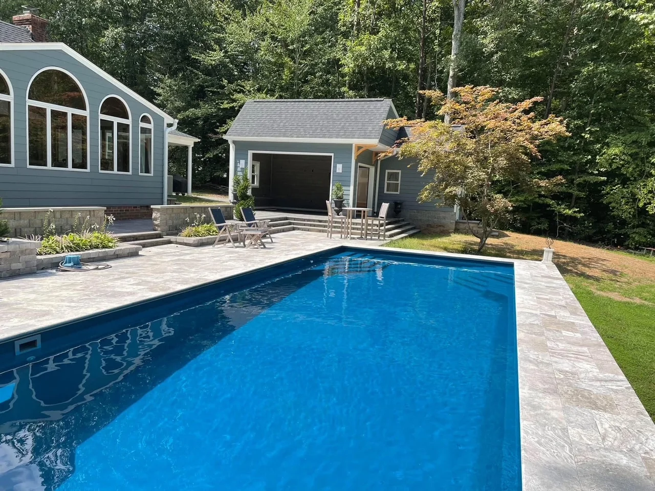 In-ground pool with blue water, gray paver deck, pool house with arched windows, and retaining walls