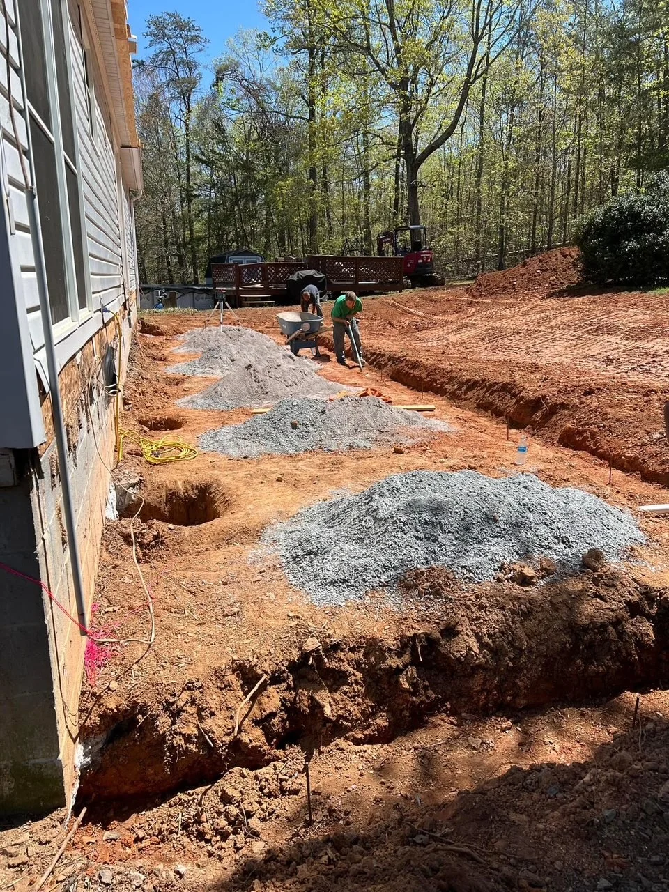 Residential foundation excavation with footer trenches, gravel base material, and site grading on wooded lot